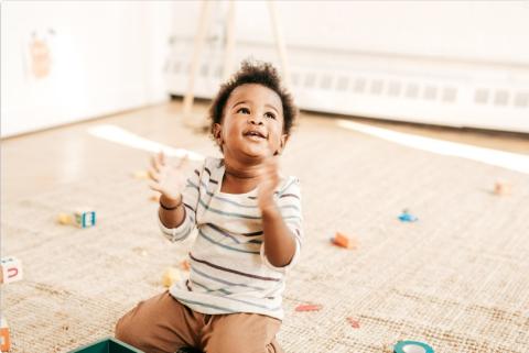 A very young child kneeling, looking up, smiling, and clapping