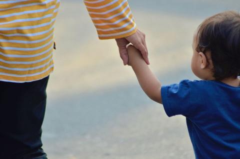 A very young child in a blue shirt holds the hand of a taller person who is wearing a yellow-and-white striped shirt.