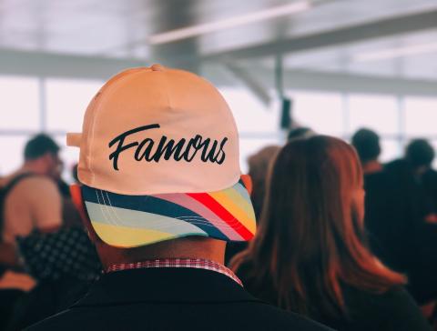 A person with their back to the camera, wearing a baseball cap that says "Famous" in cursive writing, and with a multi-colored bill.