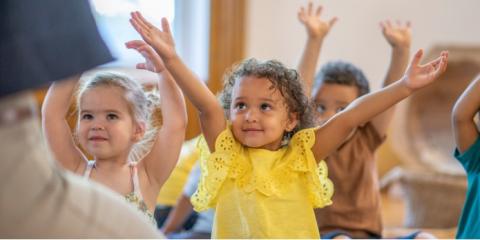 A group of toddlers sitting down with their arms stretched overhead.