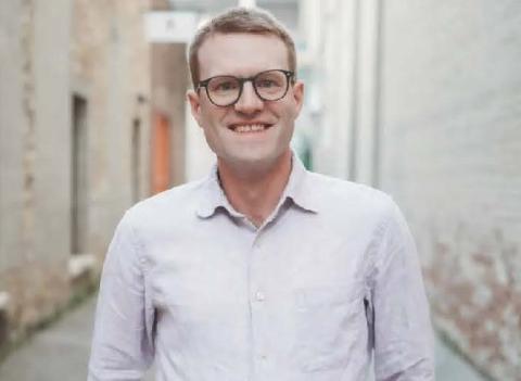 Michael T. Braun standing in an alley. He wears a light-colored oxford shirt and dark-rimmed glasses.