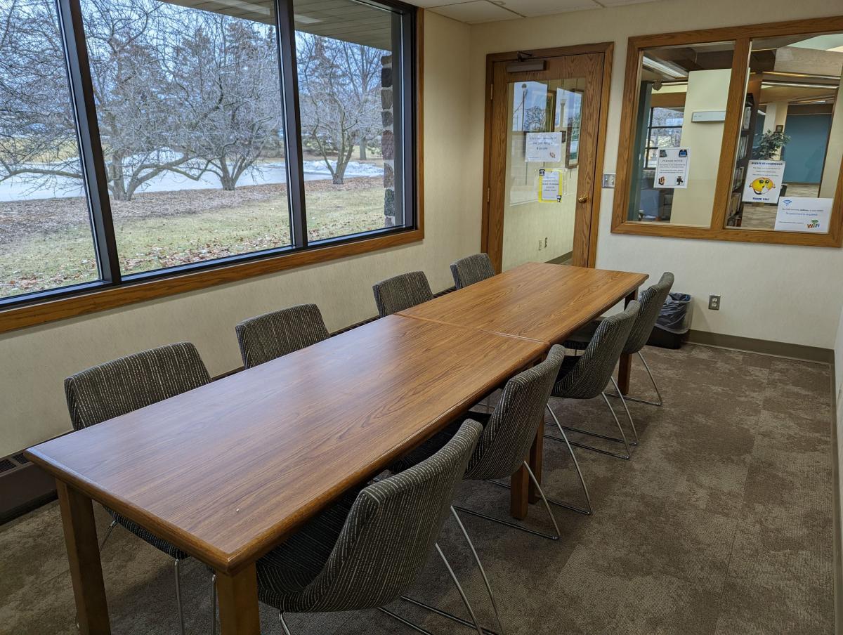 Room with windows on two sides, and a long table surrounded by eight chairs